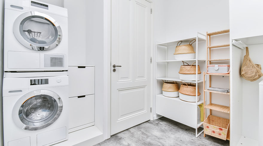 Witte wasmachine en droger staan naast een witte deur en schappen met manden in een lichte kamer.