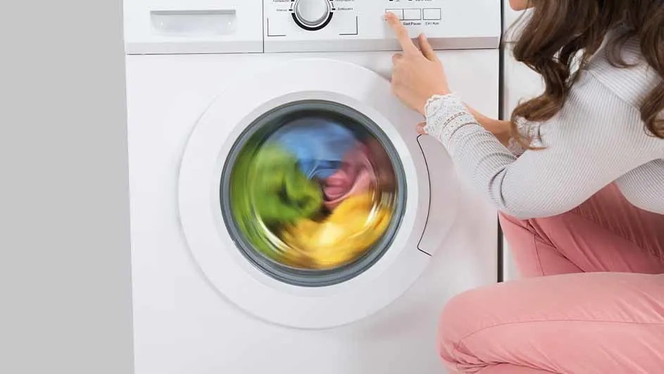 A woman starts a white washing machine filled with colourful clothes. The laundry spins quickly. No brand name is visible on the appliance.
