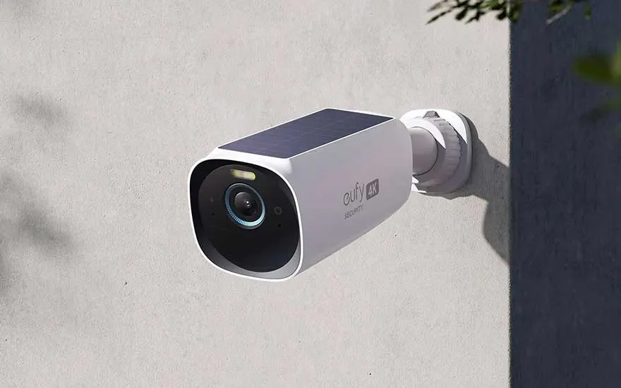 Een witte beveiligingscamera met een zonnepaneel bovenop, bevestigd aan een grijze muur met een witte draaivoet.