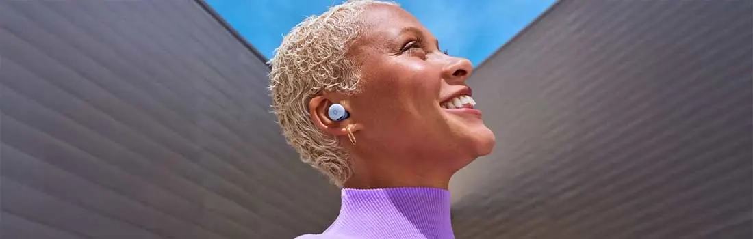 Een vrouw met kort blond haar draagt Pixel Buds en glimlacht breed, terwijl ze naar de lucht kijkt onder felle zon.