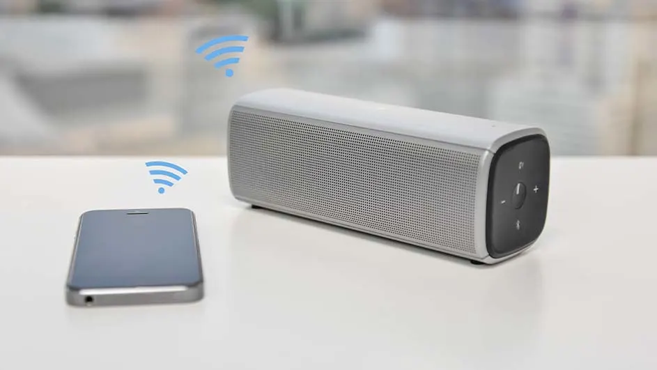 Een grijze speaker en smartphone staan op een witte tafel, met blauwe wifi-signalen erboven. De achtergrond is wazig.