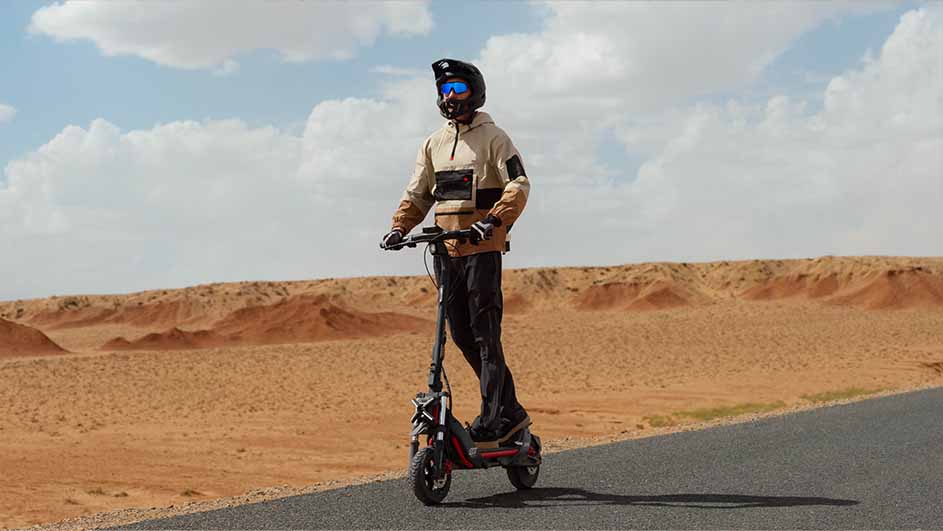 Een persoon met een helm rijdt op een elektrische scooter op een grijze weg, met zandduinen en een bewolkte lucht op de achtergrond.
