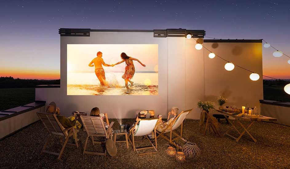A group of people are sitting outdoors in deckchairs, watching a film projected onto a wall at sunset. String lights create a warm and festive atmosphere. No brand visible.