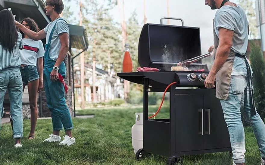 A man cooks on a gas barbecue while friends chat in the garden, creating a relaxed mood at an outdoor gathering.