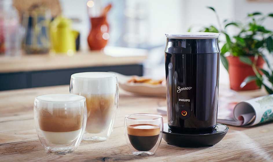 Een zwarte Senseo-melkopschuimer staat op een houten tafel, naast glazen met koffie en melkschuim, in een lichte keuken met plant.
