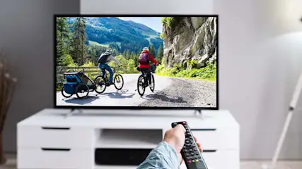 Hand met afstandsbediening voor een tv met een afbeelding van twee fietsers op een bergweg met bomen en bergen op de achtergrond.