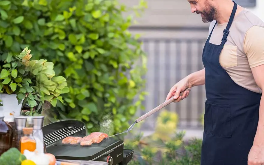 Een man met schort bakt vlees op een elektrische barbecue in een groene tuin, omringd door kruiden, olie en zomerse rust.