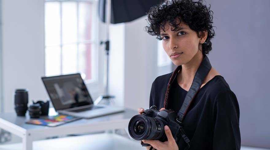 Een persoon met een zwarte camera om de nek. Een laptop en lenzen staan op een tafel in de achtergrond.