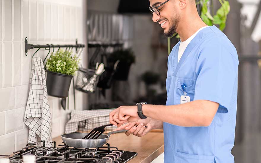 Man in blauw uniform bakt iets in een koekenpan op het gasfornuis en glimlacht terwijl hij in een nette keuken staat.
