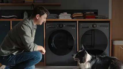 Man knielt in een nette wasruimte naast een hond, voor donkere LG wasmachines en opgevouwen wasgoed op planken.