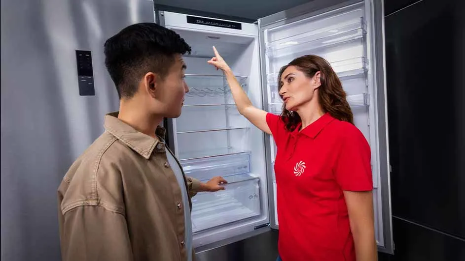 Twee mensen bij een open, zilverkleurige koelkast met witte binnenkant. Een vrouw in rood shirt wijst naar boven.