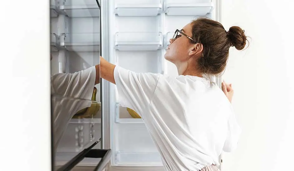 Een persoon opent een lege koelkast en kijkt naar binnen. Op de onderste plank liggen enkele bananen, de rest van de koelkast is leeg.