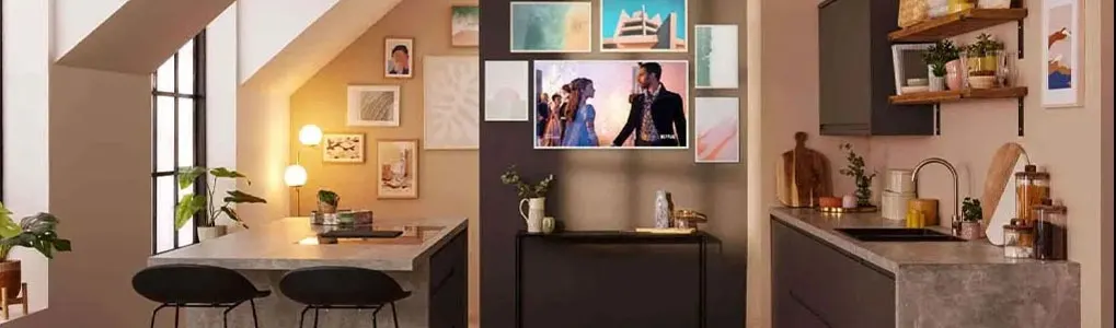 A modern kitchen with a TV showing two people, surrounded by framed art. A desk with chairs sits near a window with plants.