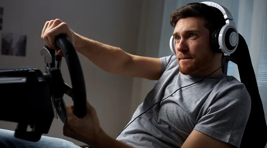 Een man met een grijze shirt en een witte koptelefoon zit achter een zwart stuur in een stoel.