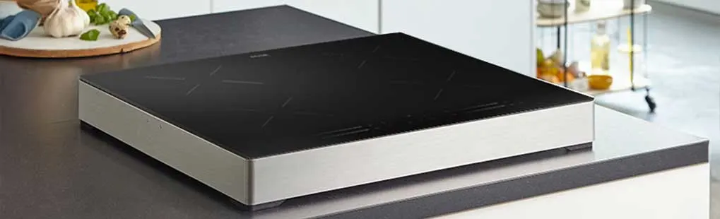 Een strakke vrijstaande inductiekookplaat met zwarte glasplaat en metalen rand staat op een moderne keukenwerkbank naast groente en knoflook.