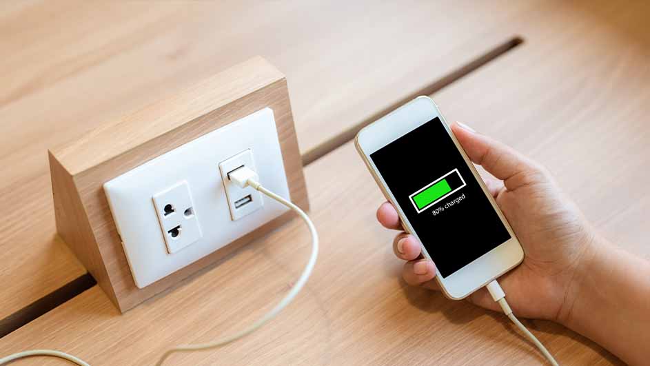 Een hand houdt een telefoon met een batterij-indicator op 80%, aangesloten op een stopcontact via een witte kabel.