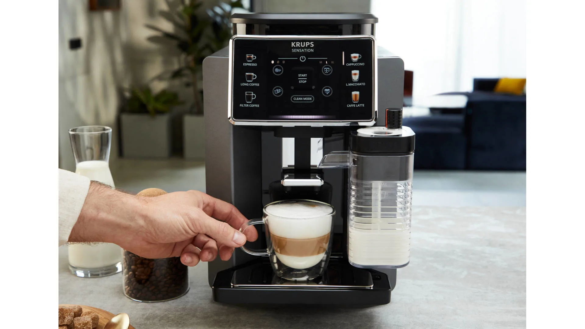 Een koffiemachine schenkt koffie in een glazen kopje. Een hand pakt het kopje. Er staat melk en koffiebonen op de achtergrond.
