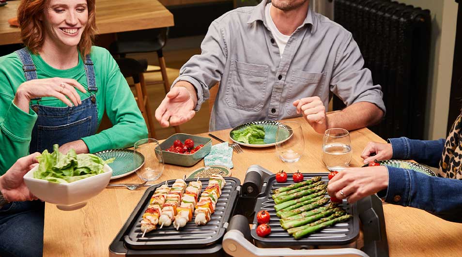 Aan een eettafel bereiden vrienden samen groentenspiesen en asperges op een tafelgrill, terwijl salades en drankjes gezellig worden gedeeld.