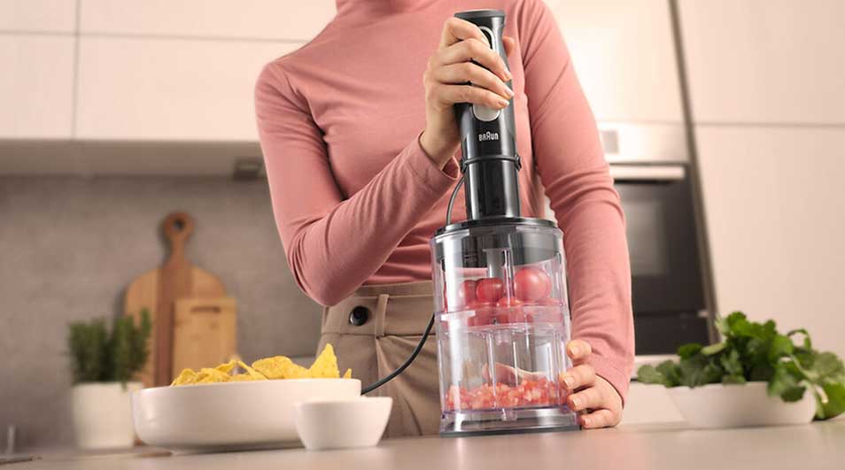 Vrouw hakt rode tomaten in een transparante hakmolen op een aanrecht, met nachochips en verse kruiden ernaast in een lichte keuken.