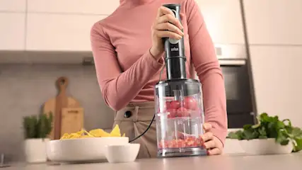 Vrouw hakt rode tomaten in een transparante hakmolen op een aanrecht, met nachochips en verse kruiden ernaast in een lichte keuken.