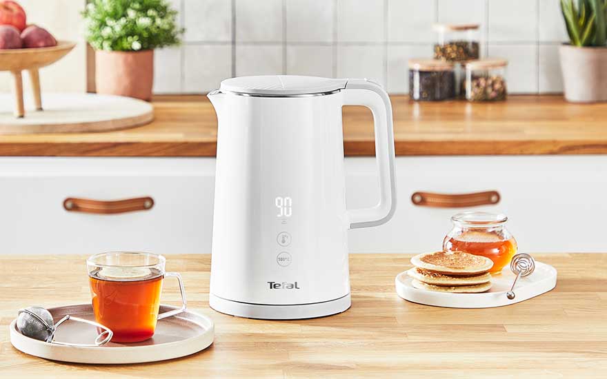 Witte Tefal-waterkoker met digitaal display staat op houten aanrecht, omringd door kop thee, pannenkoekjes et pot honing in keuken.