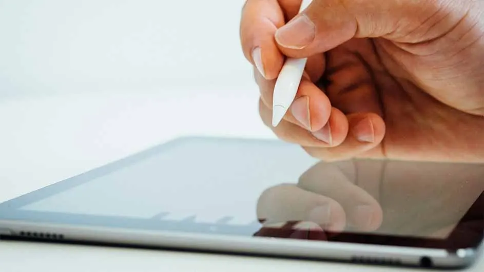A hand holds a white stylus over a grey tablet on a white surface.