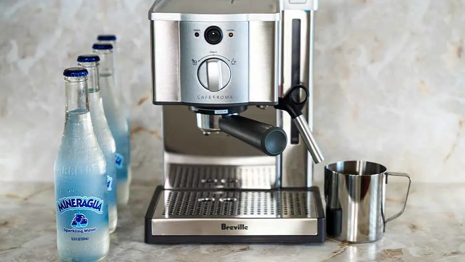 Een zilveren espressomachine staat naast drie flessen Minagua-water en een roestvrijstalen beker op een marmeren aanrecht.