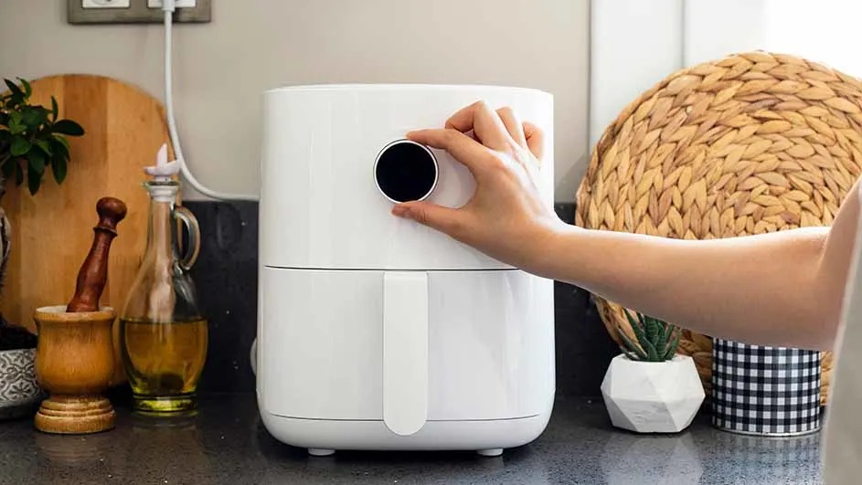 A white air fryer sits on a counter, a hand adjusts a black dial. Behind it are kitchen items: a wooden board and oil bottle.
