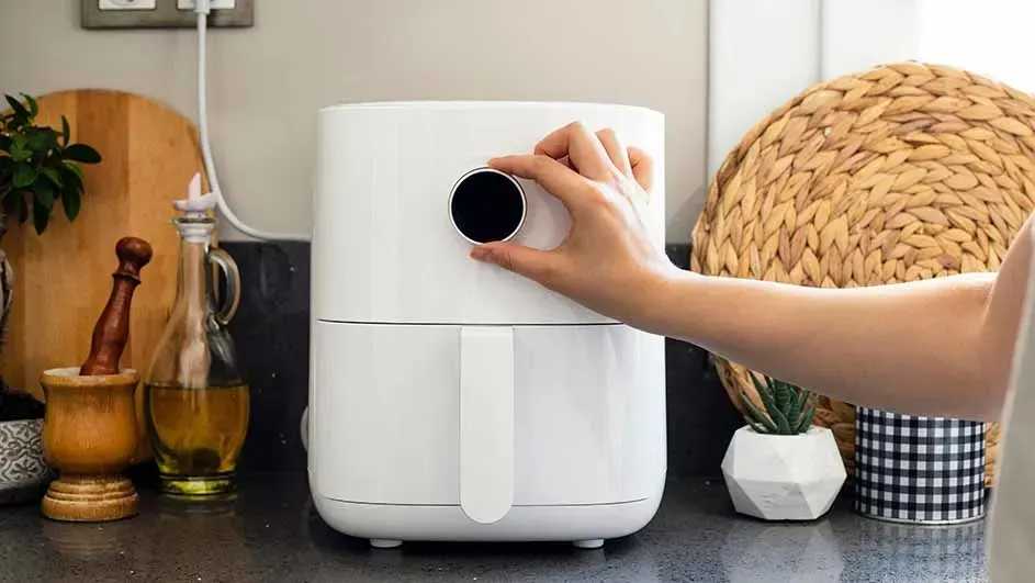A white air fryer sits on a counter, a hand adjusts a black dial. Behind it are kitchen items: a wooden board and oil bottle.