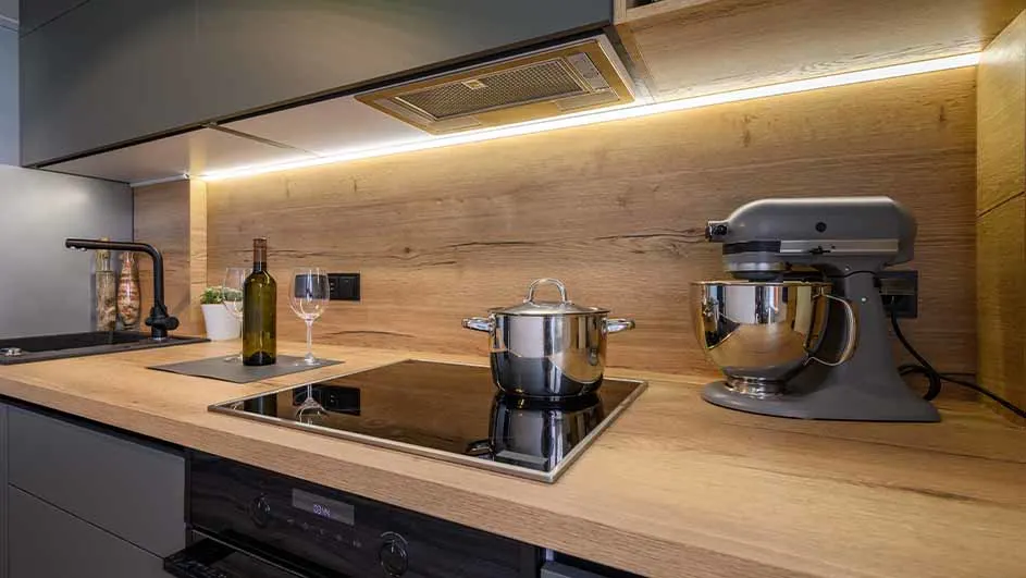 Moderne keuken met houten aanrechtblad, grijze kasten, zwarte kraan, kookplaat, zilveren pan, staafmixer en een fles wijn.