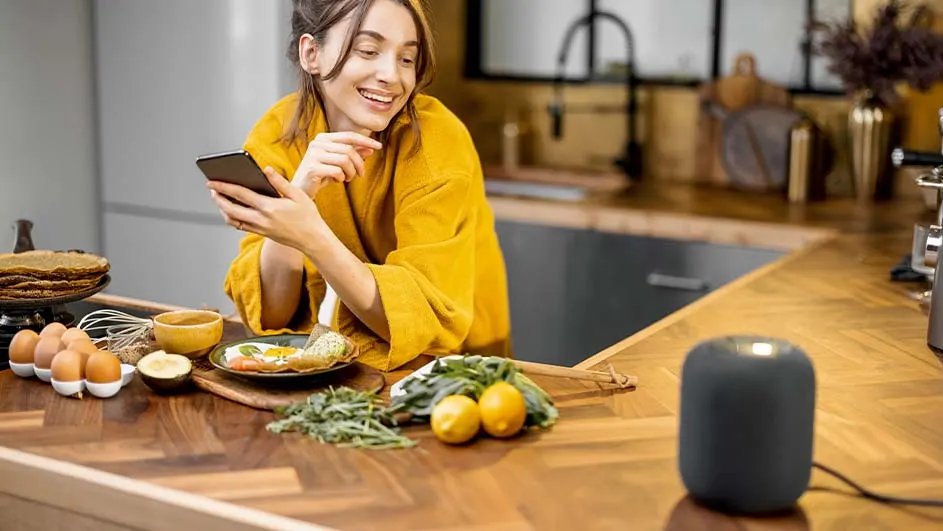 Een vrouw glimlacht naar een telefoon in een keuken. Eieren, citroenen, pannenkoeken en een grijze speaker staan op een houten aanrecht.