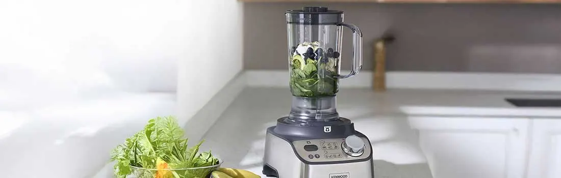 Een krachtige blender met metalen behuizing staat op een aanrecht; in de kan liggen spinazie, avocado, bosbessen en ijsblokjes voor een gezonde smoothie.