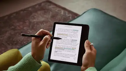 Hands hold a stylus and tablet displaying text with highlights, on a teal chair. A patterned rug is in the background.