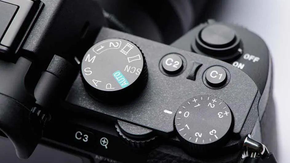 Close-up van de bovenkant van een zwarte camera met draaiknoppen voor instellingen zoals 'AUTO', 'M', 'S', 'C1' en 'C2'.