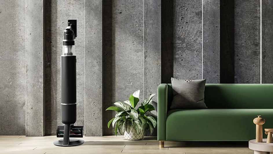 Een donkere stofzuiger staat naast een groene bank in een kamer met een betonnen muur. Een plant staat op de grond.
