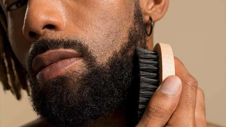 Een man borstelt zijn baard met een ronde borstel met een houten handvat en zwarte haren. Hij draagt een oorbel.