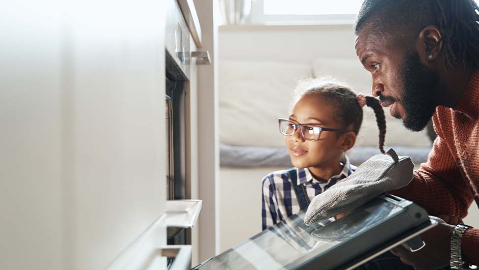 Een kind met een bril en een volwassene kijken in een oven met een grijze ovenwant.