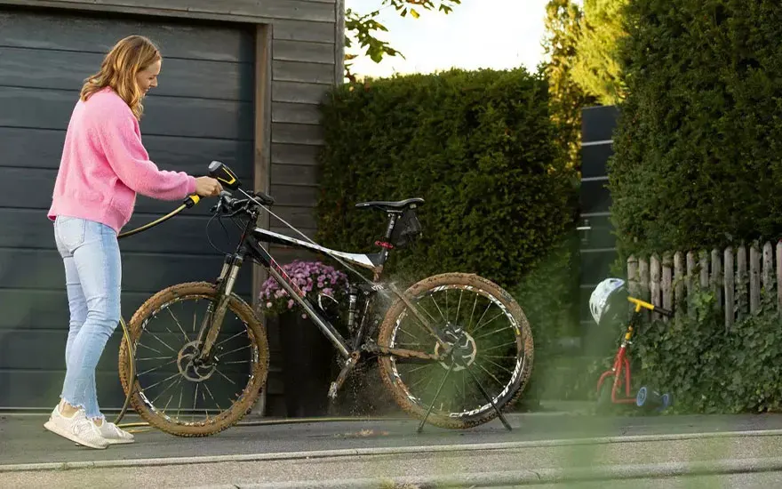 Een vrouw in roze trui reinigt haar mountainbike met een hogedrukreiniger buiten bij een grijs huis met groene planten op de achtergrond.