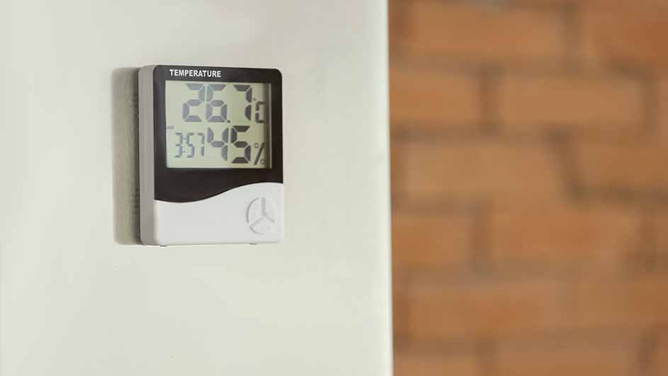 Een digitale thermometer toont 26.7 graden en 35.45% luchtvochtigheid aan een witte muur, met een bakstenen muur op de achtergrond.