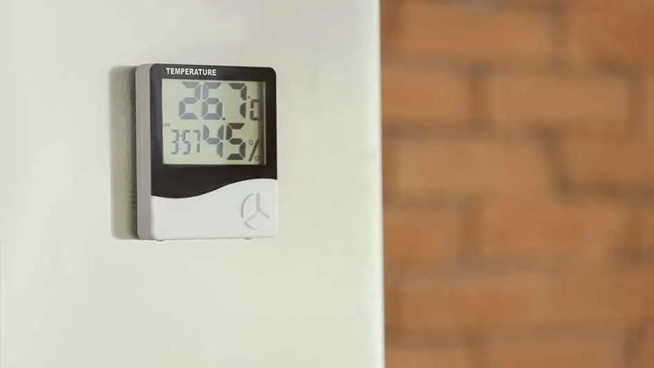 Een digitale thermometer toont 26.7 graden en 35.45% luchtvochtigheid aan een witte muur, met een bakstenen muur op de achtergrond.