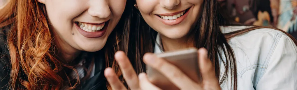 Twee jonge vrouwen lachen samen terwijl ze op een smartphone kijken; het scherm weerspiegelt hun vrolijke gezichten. Geen merk zichtbaar.