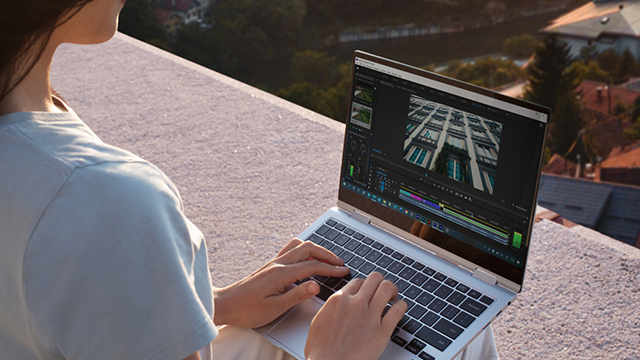Een persoon typt op een laptop met bewerkingssoftware, tegen een achtergrond van gebouwen en groen.