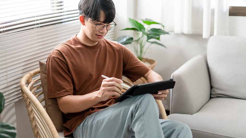 Een persoon zit in een stoel met een tablet en stylus, in een kamer met een bank en plant.