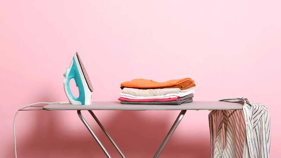 Een blauw strijkijzer staat naast een stapel kleding op een strijkplank, met een gestreept overhemd aan de zijkant tegen een roze achtergrond.