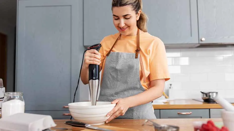 Een vrouw met een grijze schort gebruikt een staafmixer in een witte kom op een houten aanrecht in een keuken.
