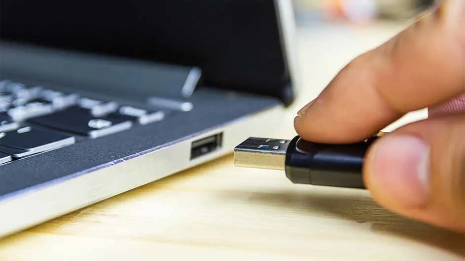 Een hand steekt een zwarte USB-stick in een zilverkleurige laptop op een houten ondergrond.