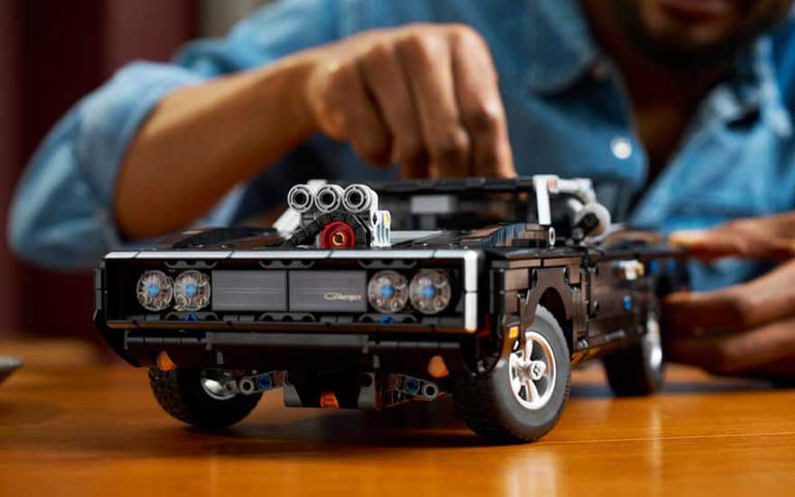 Close-up van zwarte LEGO muscle car op houten tafel, terwijl iemand achteraan kleine onderdelen zorgvuldig vastzet.
