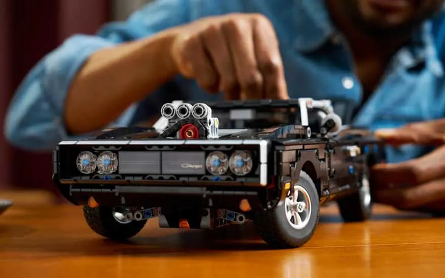 Close-up van zwarte LEGO muscle car op houten tafel, terwijl iemand achteraan kleine onderdelen zorgvuldig vastzet.