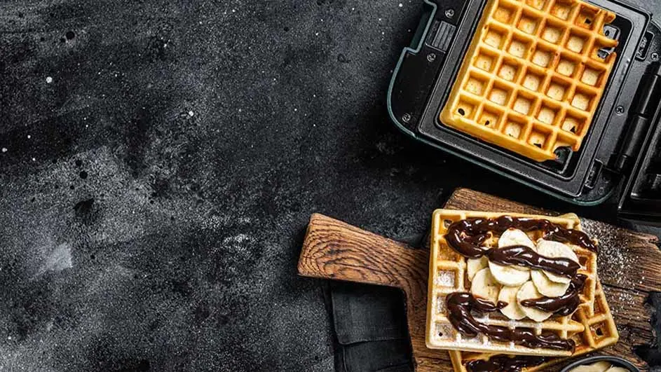 Wafels in een wafelijzer en op een houten plank met banaan en chocolade op een zwarte achtergrond.
