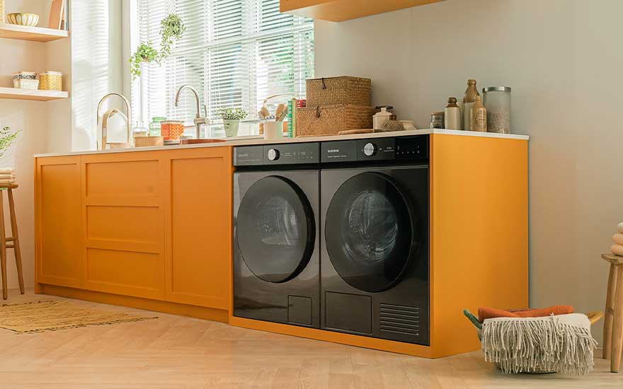 In een gezellige ruimte staan twee zwarte machines onder geel keukenblad, met planten, manden en zonlicht voor een warme sfeer.